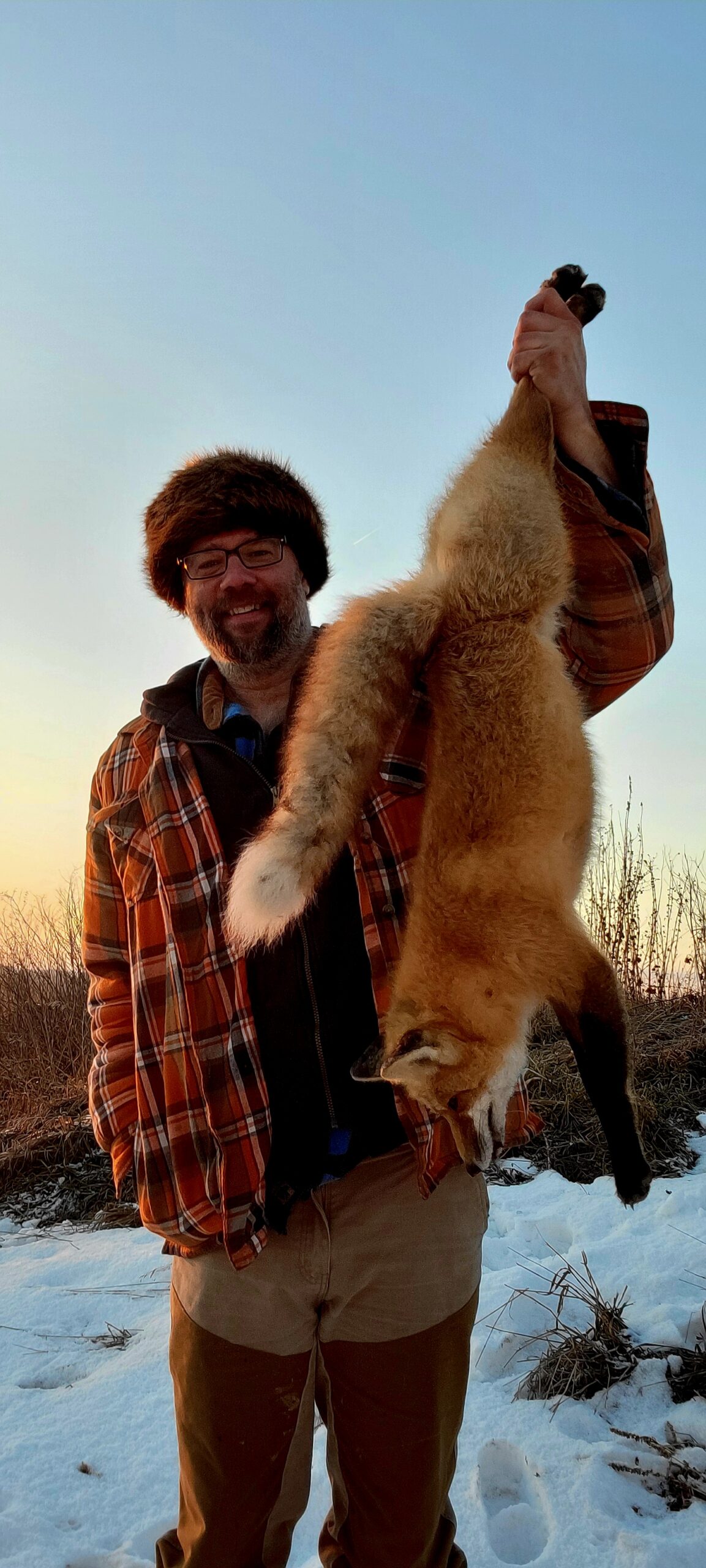Trapper holding a red fox after fresh snowfall at sunrise.